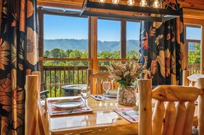 A dining table set for two with a view of a forested landscape through large windows. - Firefly Forest • Stunning Mtn Views • Hot Tub • Sauna • Firepit • 5 Min to Pkwy! (Pigeon Forge)