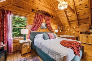A wooden cabin bedroom with a bed, bedside tables, a window with curtains, a chandelier, and a view of greenery outside.