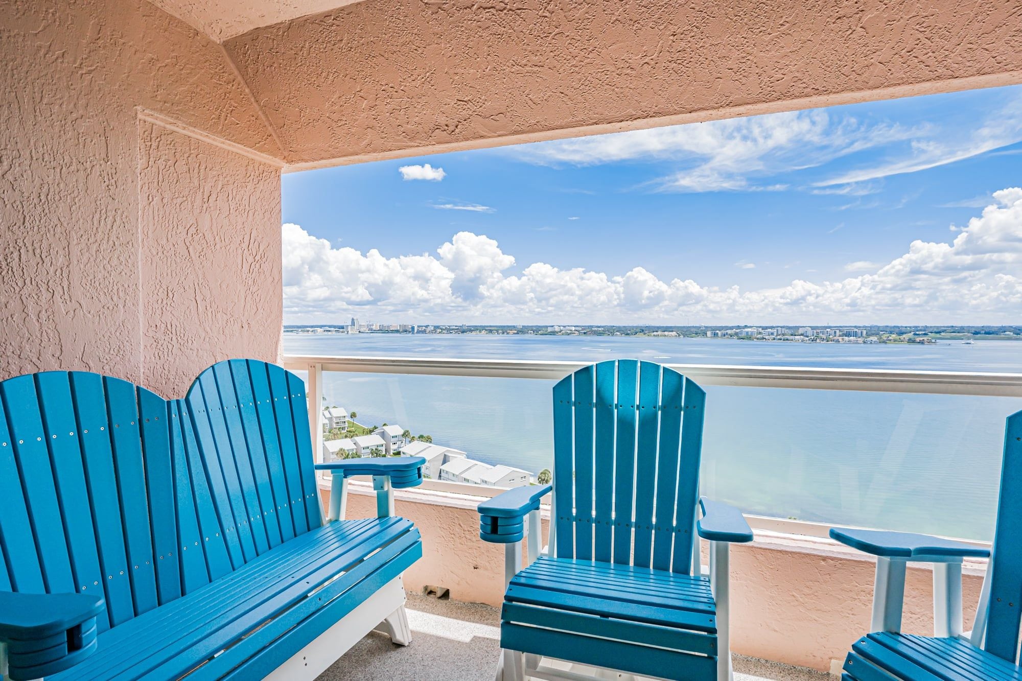 A balcony with two blue Adirondack chairs and a view of a body of water and distant cityscape.
