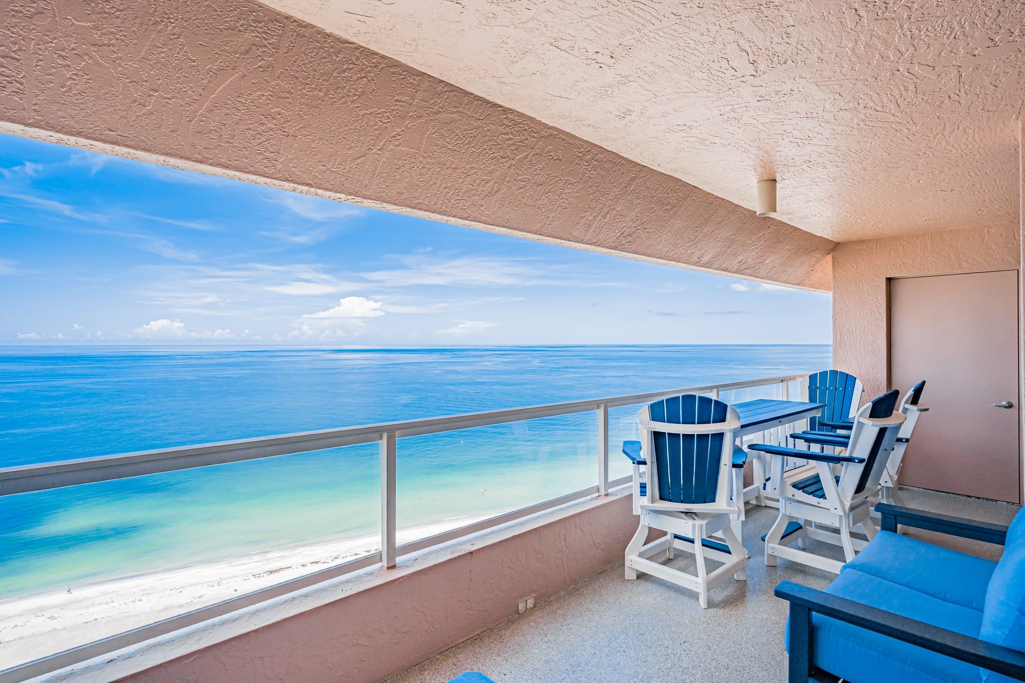 A balcony with a view of the ocean, a table, and blue chairs.