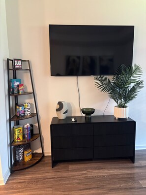 A living room with a black entertainment unit, a flat-screen TV mounted on the wall, a small potted plant, and a game shelf.