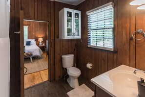 A wooden-paneled bathroom with a toilet, sink, and window with blinds. - 1929 Creek House at HollyOaks on the Marsh (Savannah)