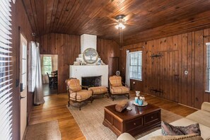 A wooden-paneled room with a fireplace, two armchairs, a coffee table, and a ceiling fan. - 1929 Creek House at HollyOaks on the Marsh (Savannah)