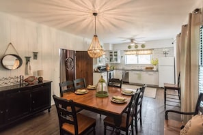 A dining area with a wooden table and chairs, a hanging light fixture, and a view into the kitchen. - 1929 Creek House at HollyOaks on the Marsh (Savannah)