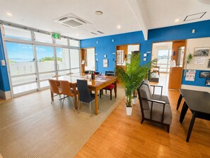 A hotel room with a large window, a wooden table, chairs, a potted plant, and a bed.