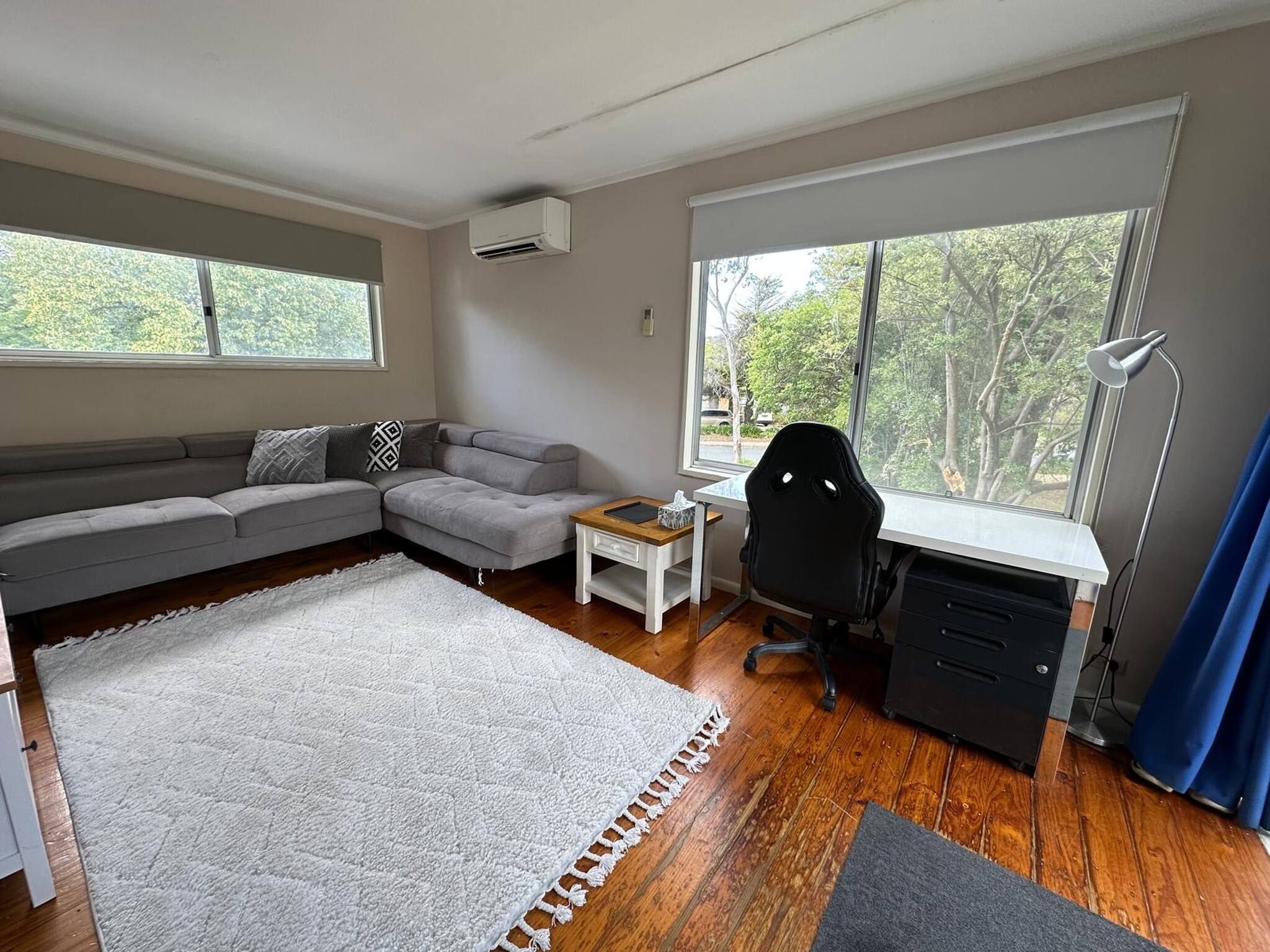 Uma sala de estar moderna com um sofá seccional, uma mesa pequena, uma escrivaninha com cadeira e grandes janelas com vista para árvores.