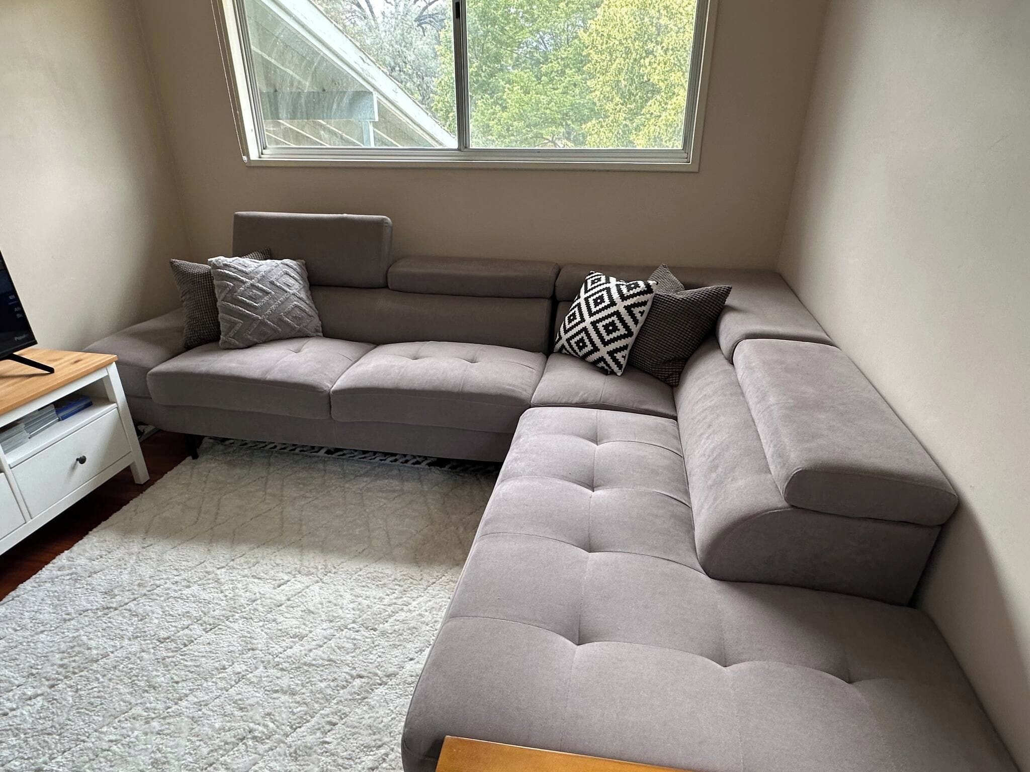Una sala moderna con un sofá modular, un tocador blanco pequeño y un ventanal con vistas a árboles.