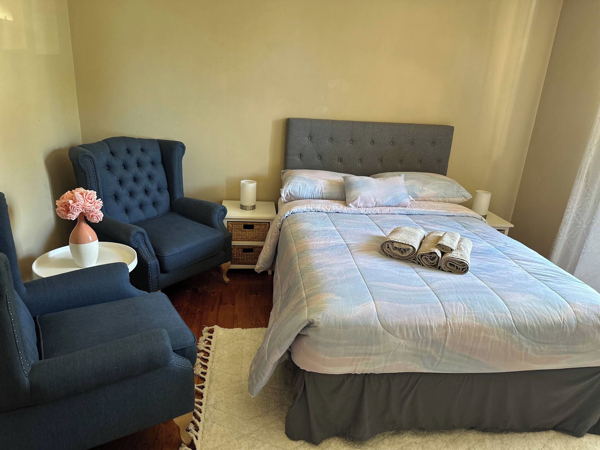 Un dormitorio con una cama, dos sillas, una mesita de noche y un florero con flores.