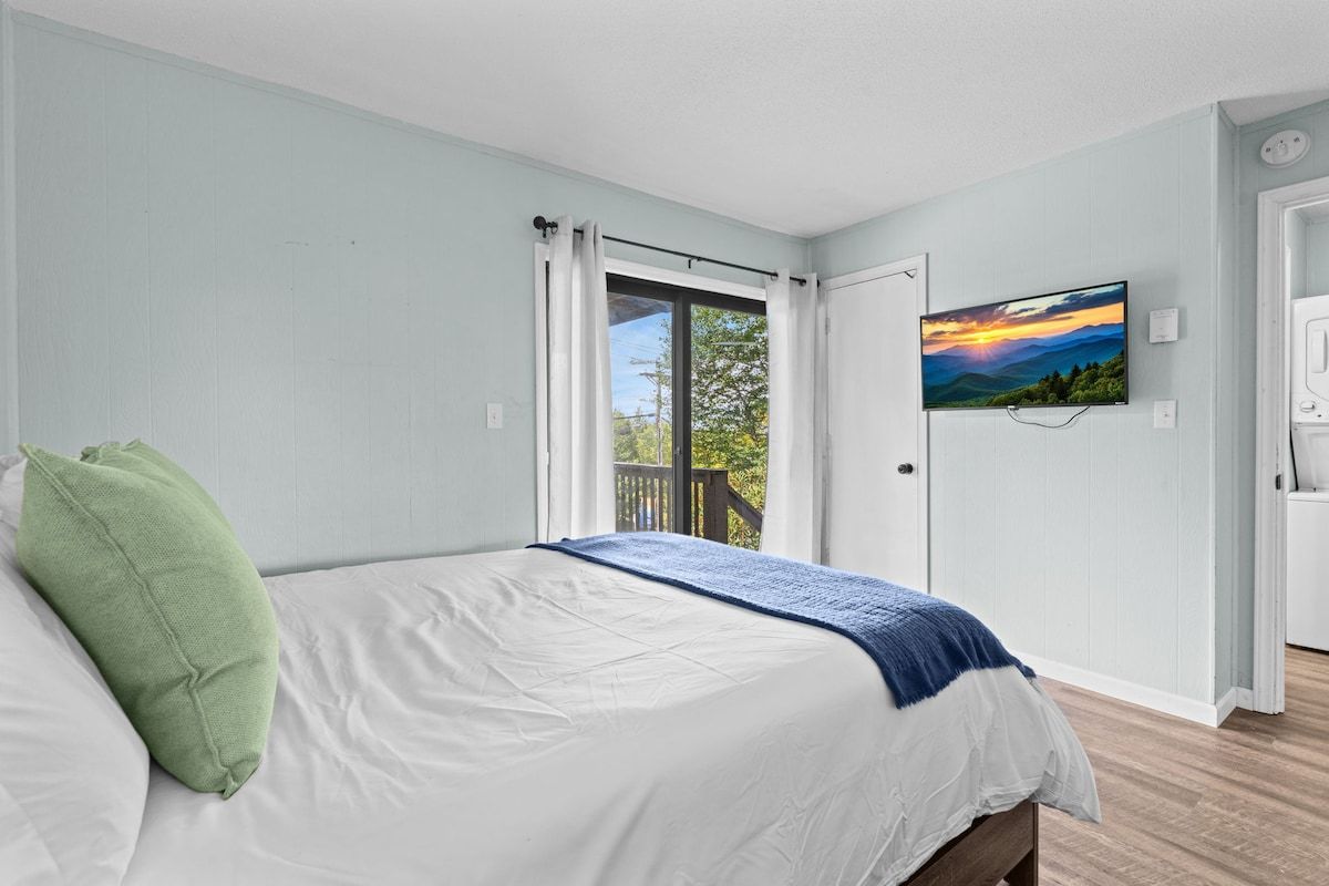 A bedroom with a bed, a window with curtains, a television on the wall, and a door leading to another room.