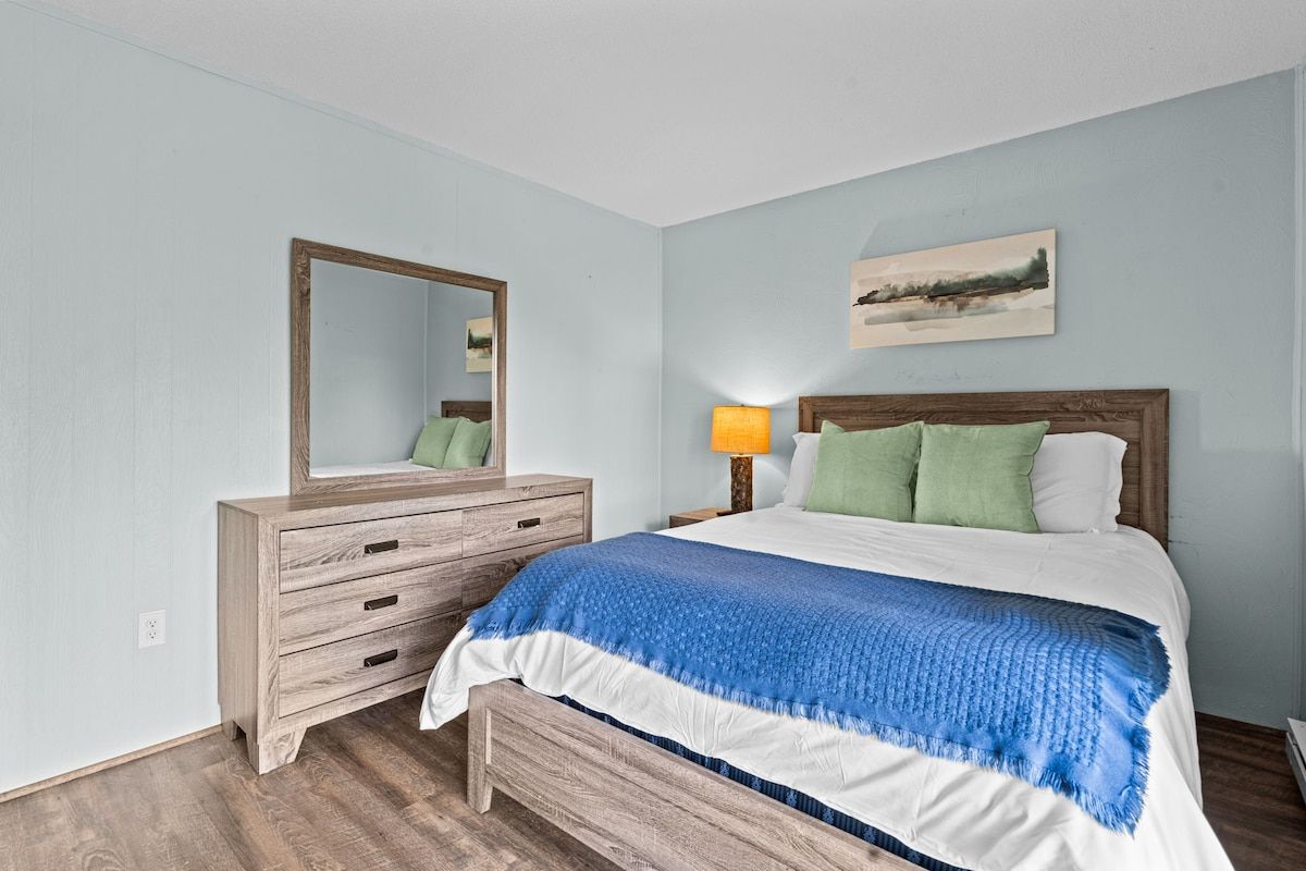 A bedroom with a wooden dresser, a bed with a blue and white blanket, a nightstand with a lamp, and a wall-mounted mirror.