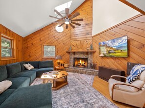 A cozy living room with a stone fireplace, wooden walls, a ceiling fan, and a television mounted on the wall. - Cozy High Country Cabin Near Banner Elk (Banner Elk)