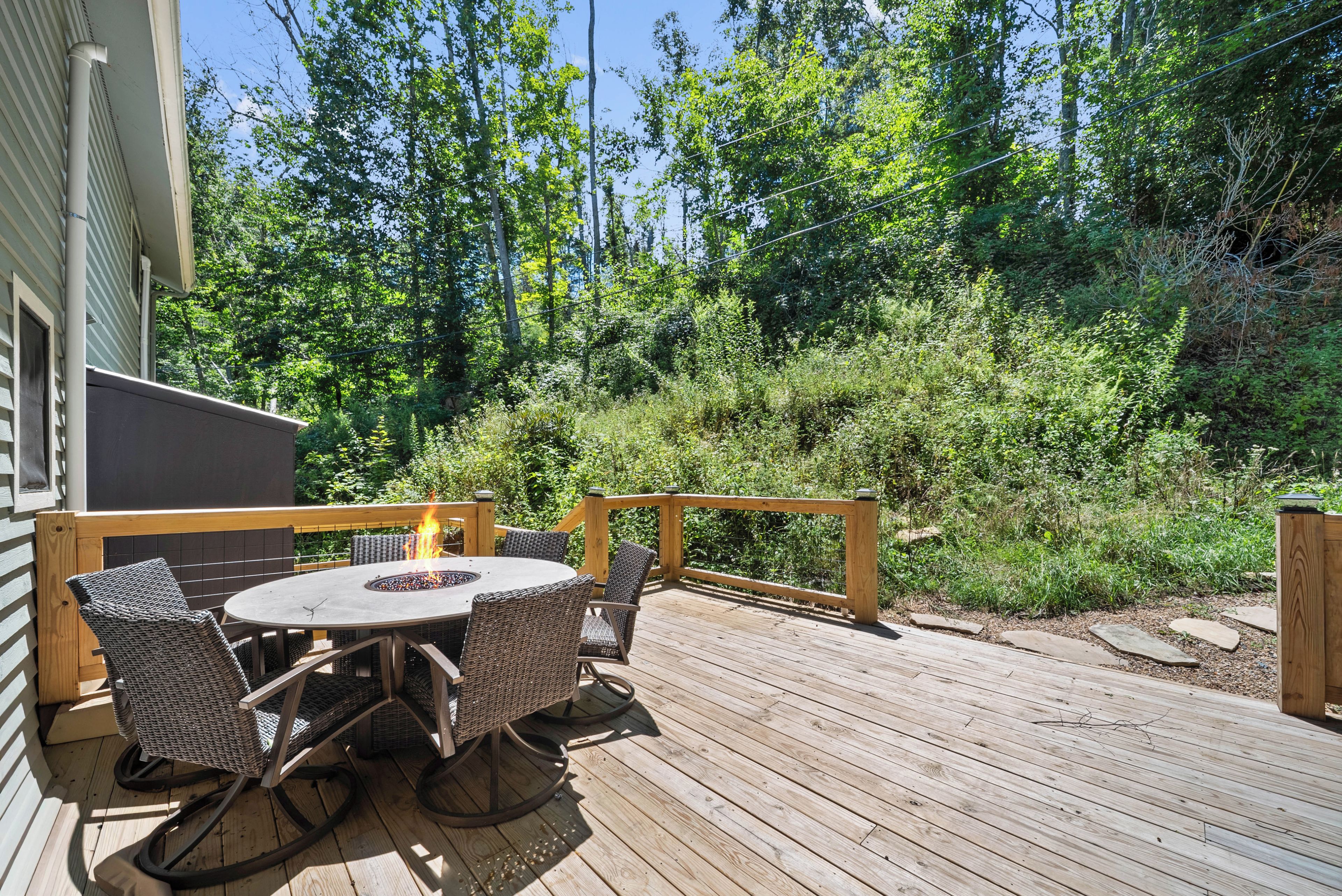 A wooden deck with a round table and wicker chairs, surrounded by greenery.