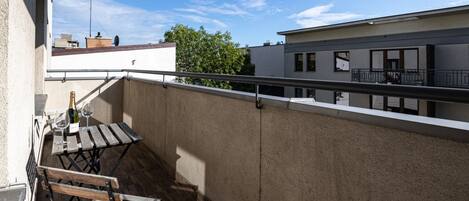 A balcony with a wooden table and chairs, a bottle of wine, and a view of neighboring buildings and trees.