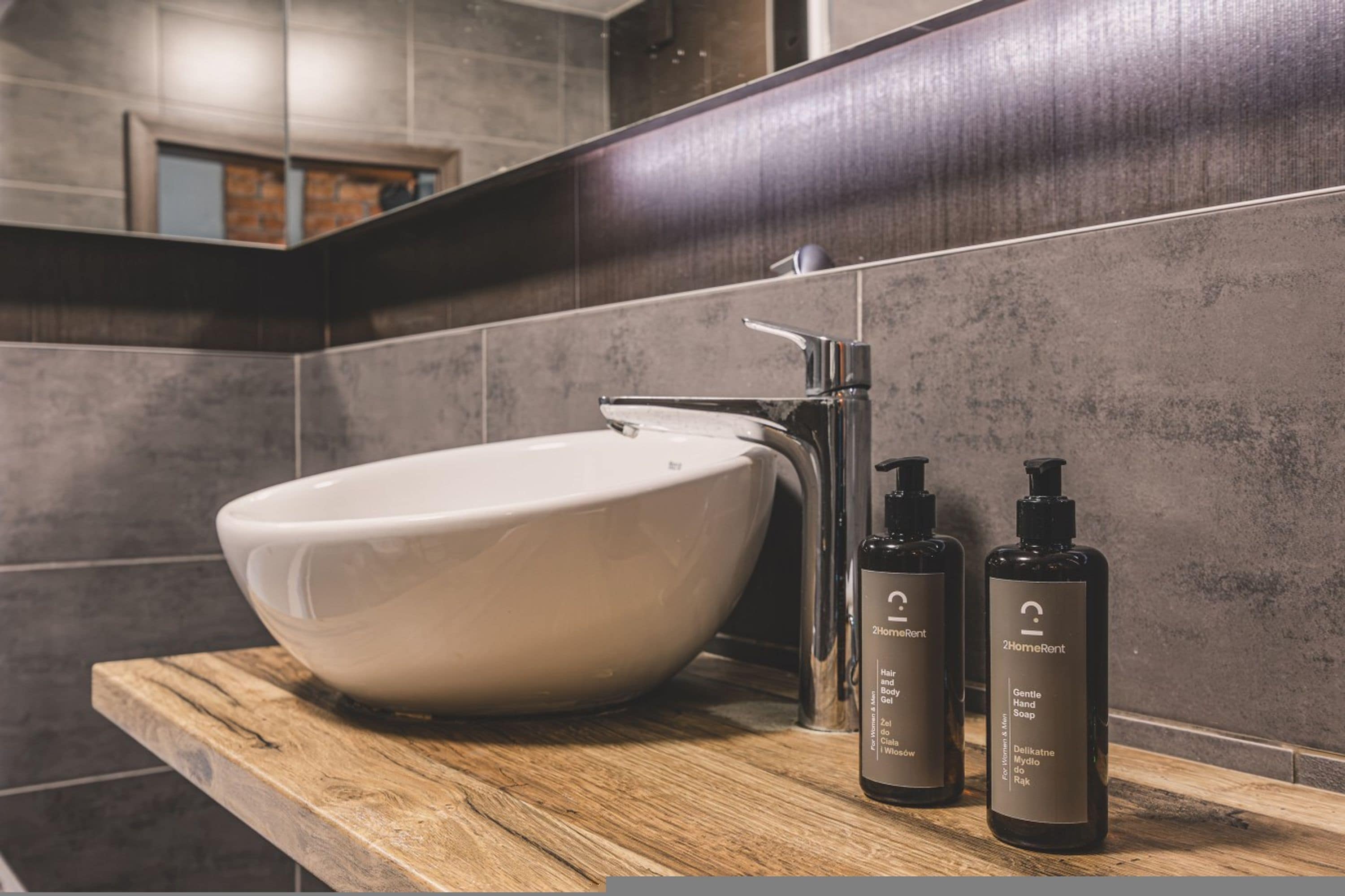 A modern bathroom with a large white sink, a chrome faucet, and two bottles of 2HomeRent hair and body gel on a wooden countertop.