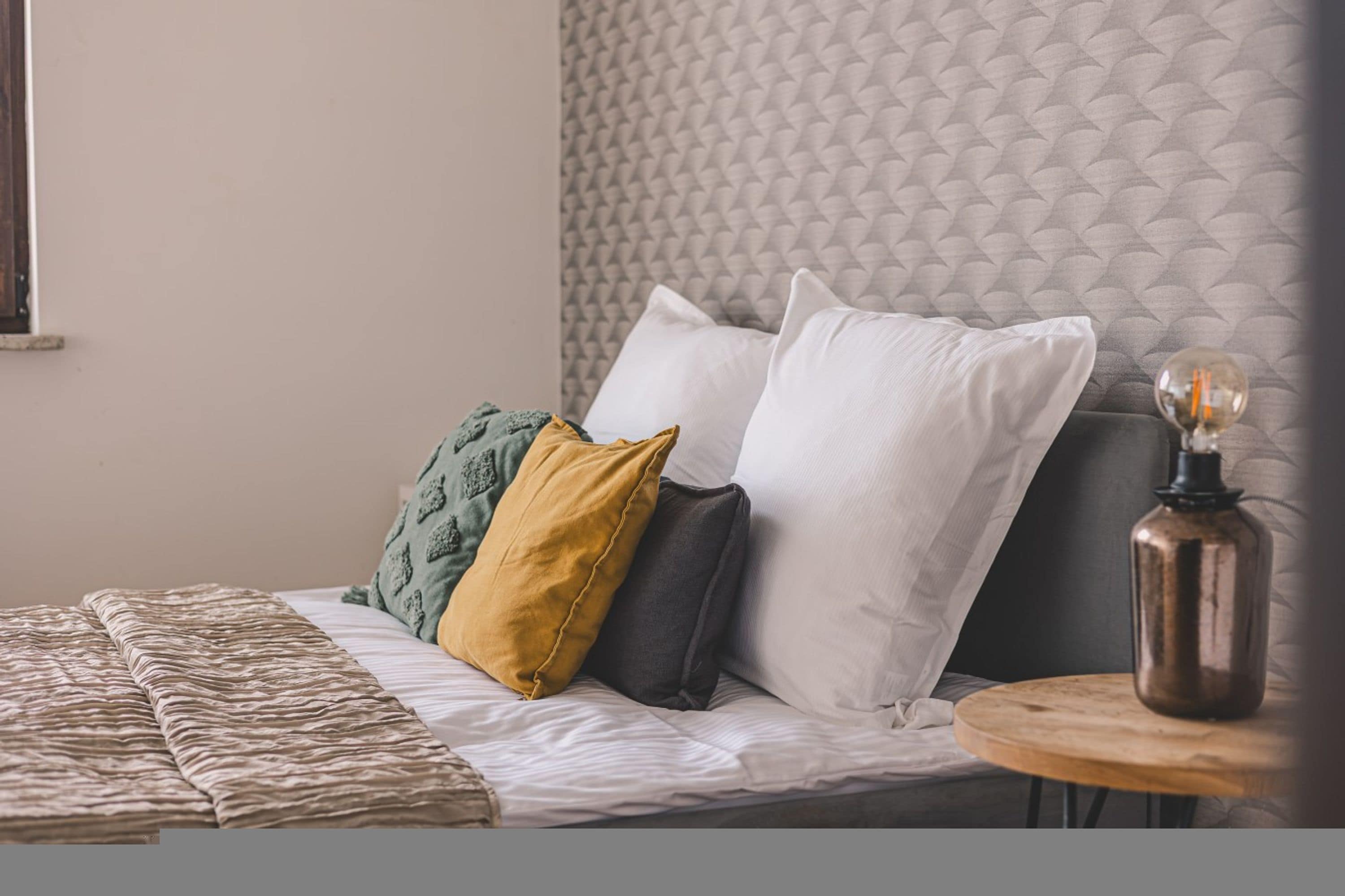 Una cama con cabecera texturizada, varios almohadones, una colcha estampada y una mesita de noche con lámpara.