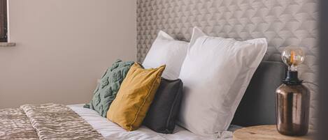 Una cama con cabecera texturizada, varios almohadones, una colcha estampada y una mesita de noche con lámpara.