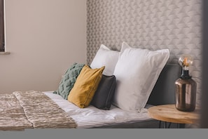 Ein Bett mit strukturierter Kopfteil, mehreren Kissen, einer gemusterten Decke und einem Nachttisch mit einer Lampe.