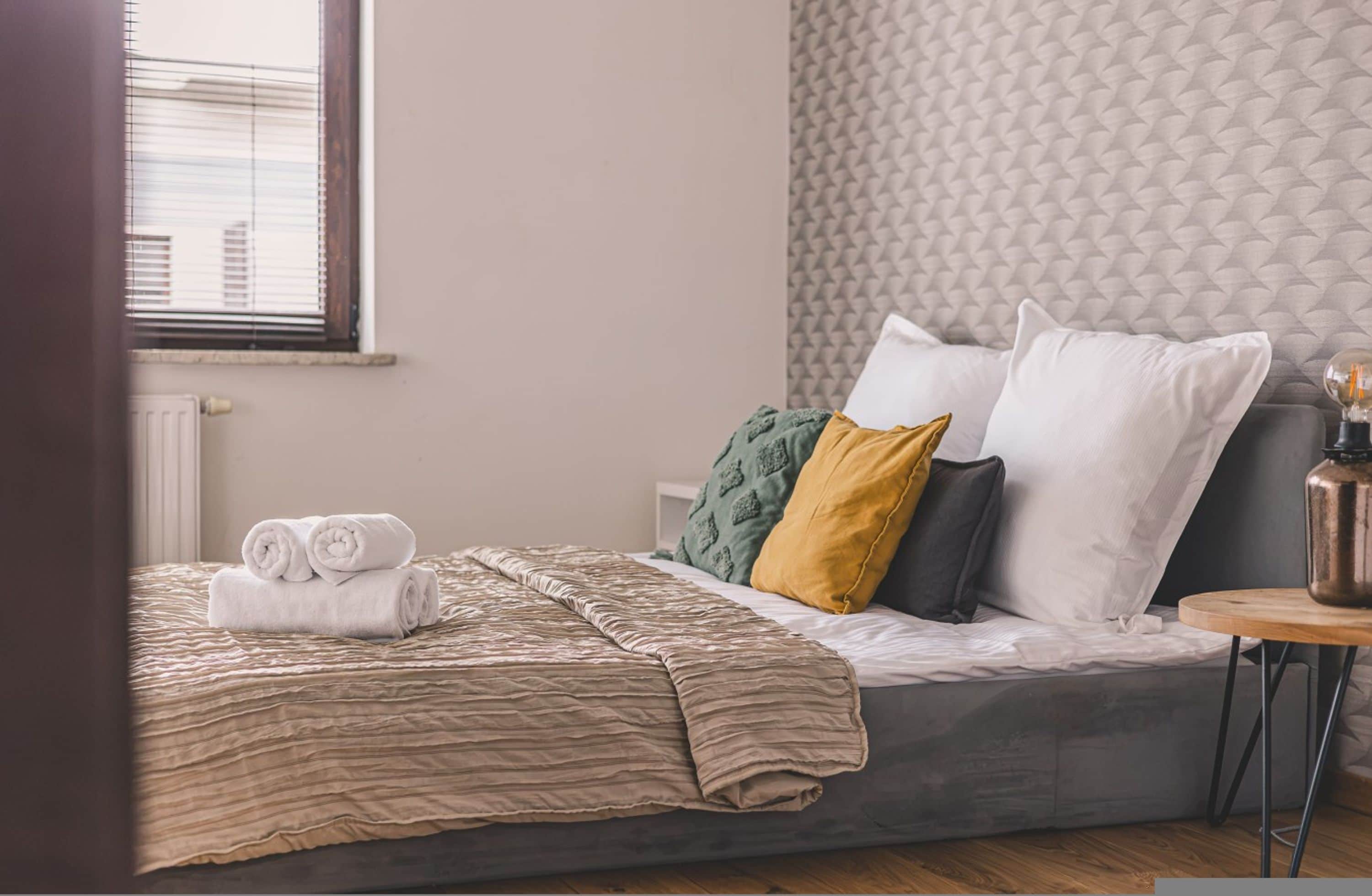 Een keurig opgemaakt bed met opgerolde handdoeken, een behang met patroon, en een klein nachtkastje met een lamp.