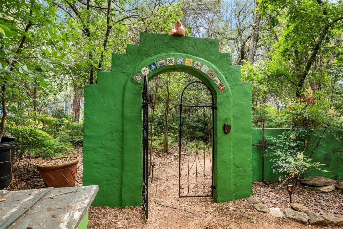 Une arche verte ornée d’un motif décoratif, qui mène à une zone boisée où se trouvent un banc en bois et des plantes en pot.
