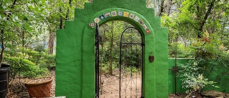 Une arche verte ornée d’un motif décoratif, qui mène à une zone boisée où se trouvent un banc en bois et des plantes en pot.