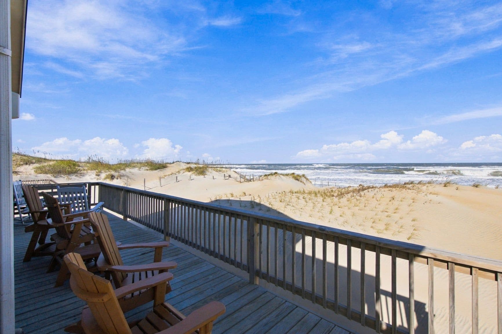 A wooden deck with chairs overlooking a beach with dunes and the ocean.