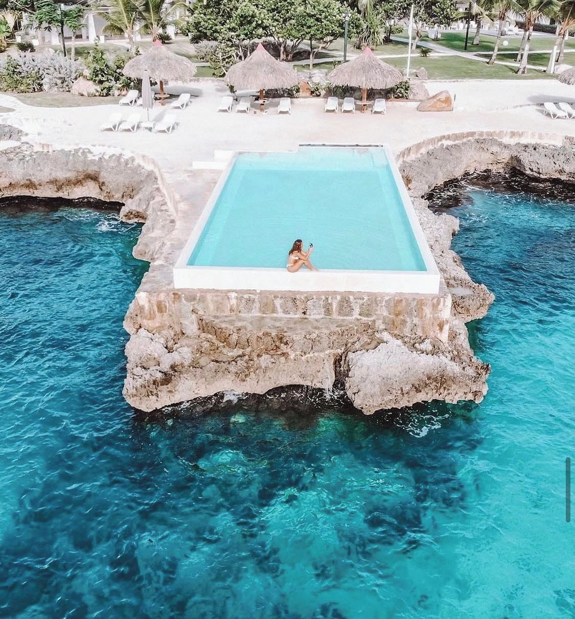 A rectangular pool with a person sitting inside, surrounded by white lounge chairs and thatched umbrellas, set against a backdrop of greenery and a clear blue sky.