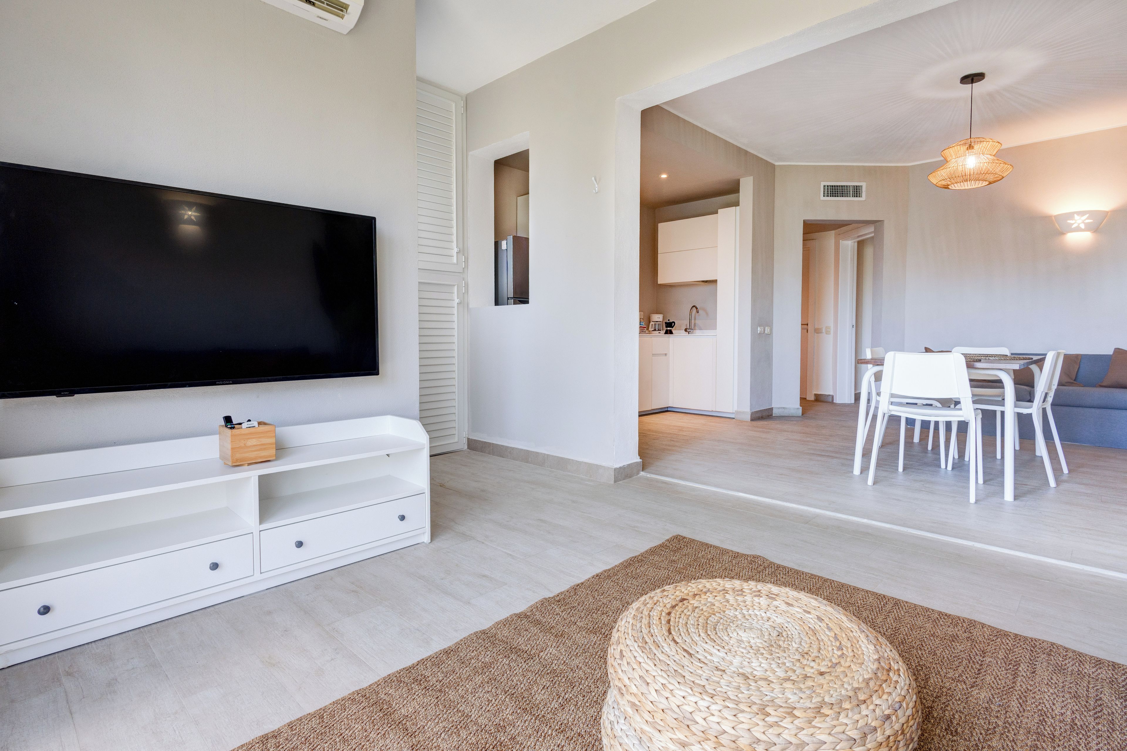 Un salon moderne avec un téléviseur à écran plat, des meubles blancs, un pouf en osier et une cuisine en arrière-plan.