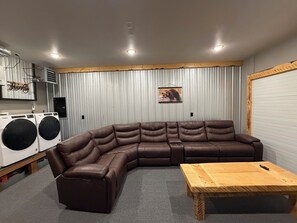 Une pièce comprenant un canapé d’angle, une table basse en bois, un lave-linge et une grande fenêtre avec des rideaux.