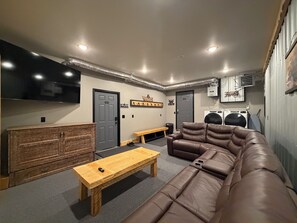 A room with a brown leather sofa, a wooden bench, and a small table. There are two doors, one labeled 'WELCOME' and another with a sign above it. A washing machine is visible in the background. - Spacious 3-bedroom 2 living apartment with Alaska theme. TV’s in every room. (North Pole)