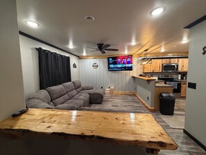 A modern living room with a sectional sofa, a wooden coffee table, a ceiling fan, a TV mounted on a wall, and a kitchen area with wooden cabinets and appliances. - Spacious 3-bedroom 2 living apartment with Alaska theme. TV’s in every room. (North Pole)