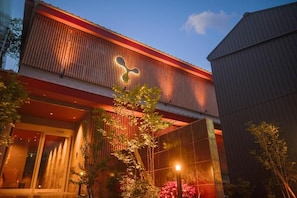 A building with a prominent logo on the facade, a glass entrance, and a well-lit outdoor area with plants. - Amu  (Beppu)