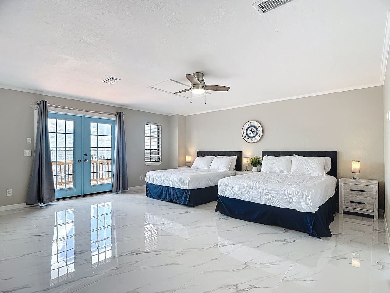 A bedroom with two beds, a ceiling fan, a clock, and a large window with curtains.