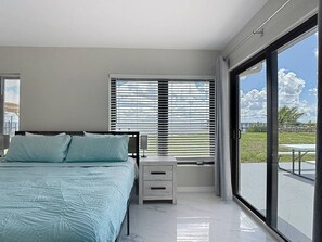 A bedroom with a bed, a nightstand, a lamp, and a view of the outdoors through large windows. - Northshore Unobstructed View 5BR 4BA Sleep 16 (Port Charlotte)