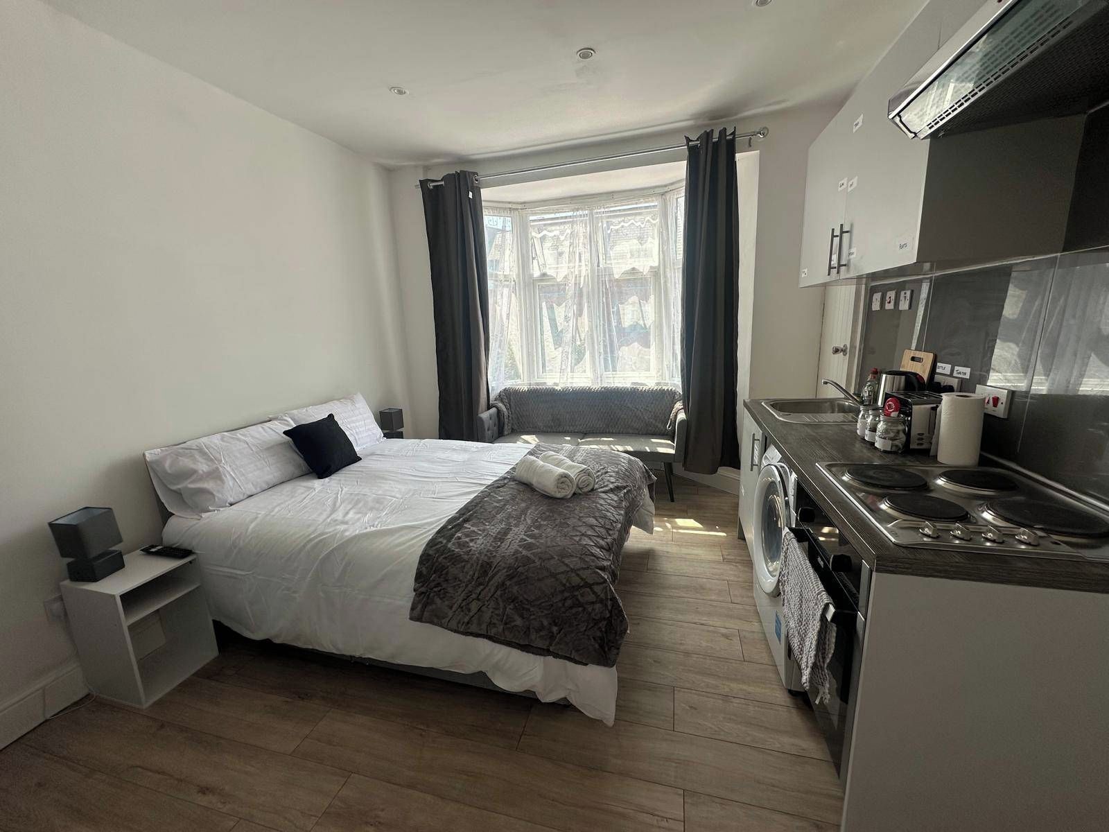 A hotel room with a bed, a sofa, a kitchenette, and a window with curtains.