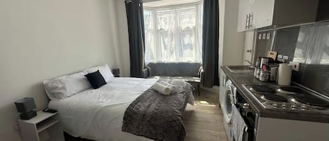 A hotel room with a bed, a sofa, a kitchenette, and a window with curtains.
