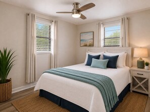 A bedroom with a bed, a ceiling fan, a nightstand with a lamp, and a window with curtains. - Shark Tooth Coastal Retreat (Englewood)