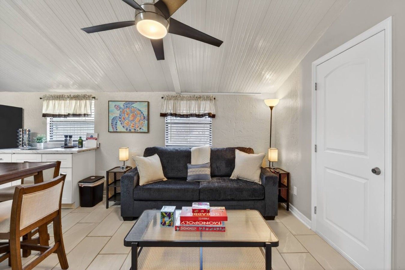 A living room with a sofa, two side tables, a coffee table with board games, a ceiling fan, and a dining area with a table and chairs.