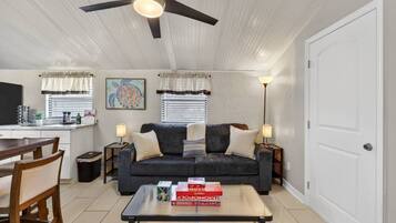A living room with a sofa, two side tables, a coffee table with board games, a ceiling fan, and a dining area with a table and chairs.