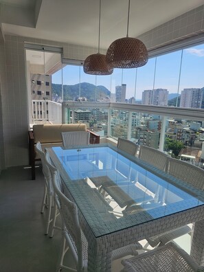 A balcony with a glass-top dining table, wicker chairs, and a view of the cityscape. - Beautiful Luxury Resort Apartment (Guarujá)