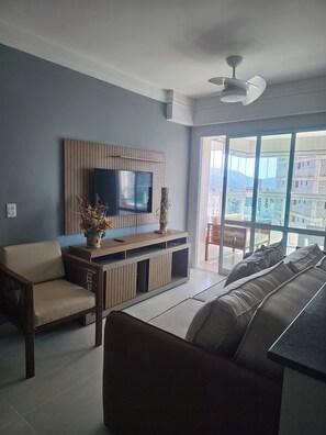 A modern living room with a flat-screen TV, a wooden entertainment unit, a sofa, and a balcony with a view of buildings. - Beautiful Luxury Resort Apartment (Guarujá)