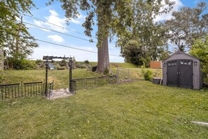 Une zone herbeuse entourée d’une clôture métallique, agrémentée d’une pergola en bois et d’un abri de jardin.