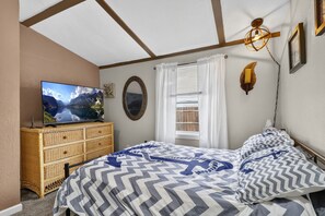 Une chambre à coucher avec un lit, un téléviseur posé sur une commode, une fenêtre avec des rideaux et un luminaire fixé au mur.