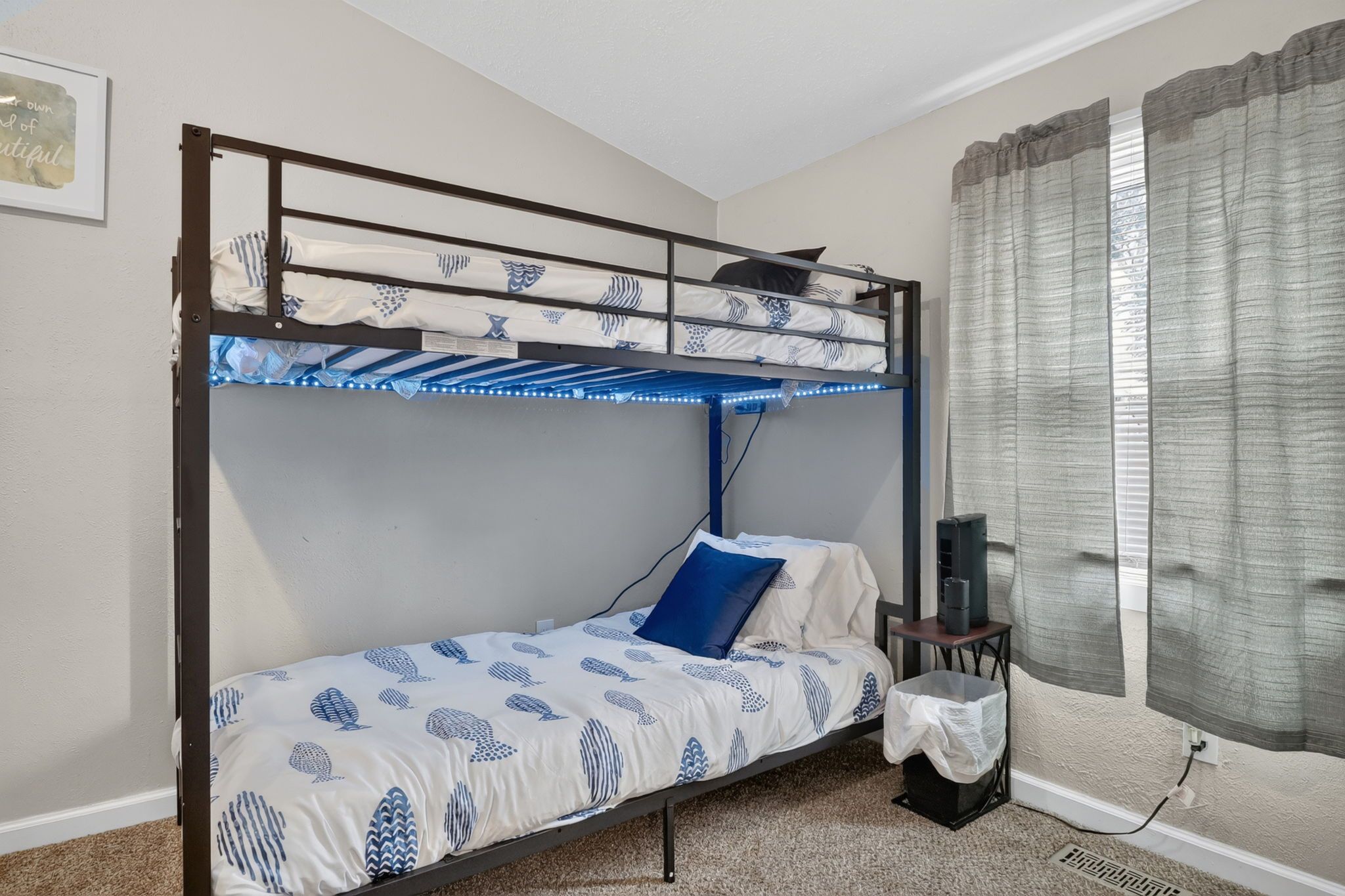 A bunk bed with a patterned bedspread, a blue pillow, a small table with a speaker, and a window with curtains.