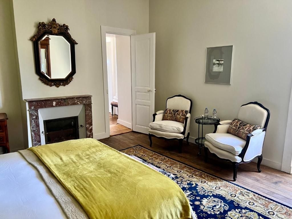Une chambre à coucher avec un lit, une cheminée, deux fauteuils, une petite table et un miroir.
