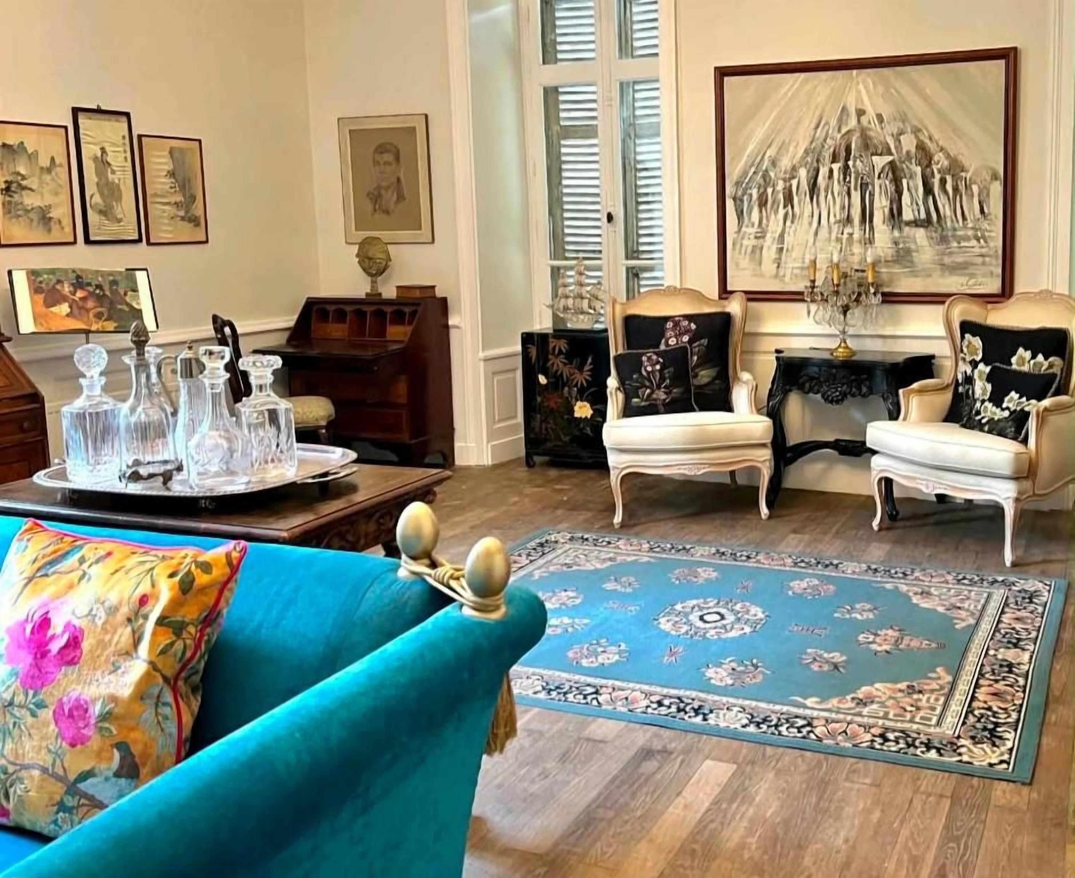 Une pièce avec un tapis bleu à motifs, deux fauteuils blancs, un canapé turquoise et un lustre.