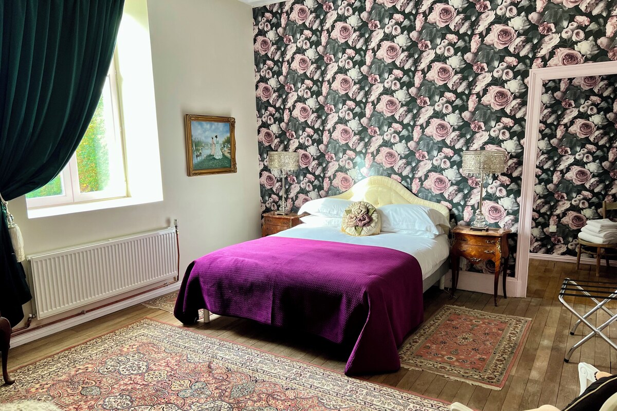 Un dormitorio con cama, mesita de noche, lámpara, silla, radiador y ventana con cortinas.