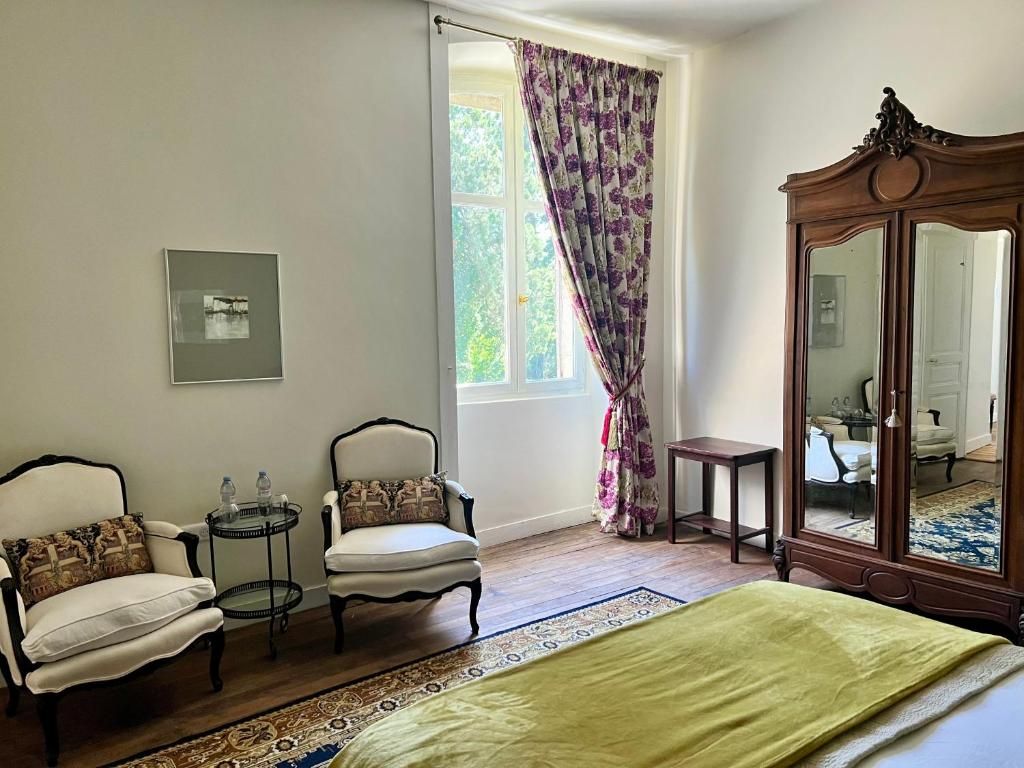 Un dormitorio con una cama, dos sillones, una mesita, un espejo y una ventana con cortinas.