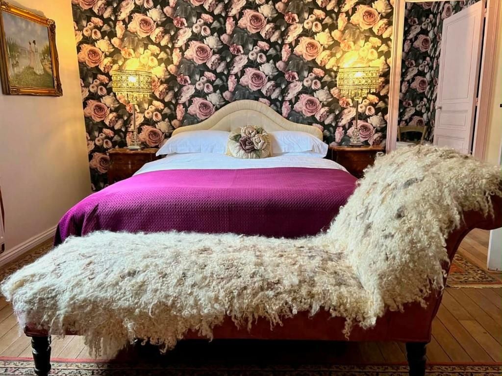 Een bed met een paars deken en een witte, pluizige deken, een muur met bloemenbehang en twee wandlampen.