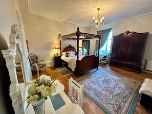 A four-poster bed with a canopy, a wooden wardrobe, a desk with a vase of flowers, and a chair.