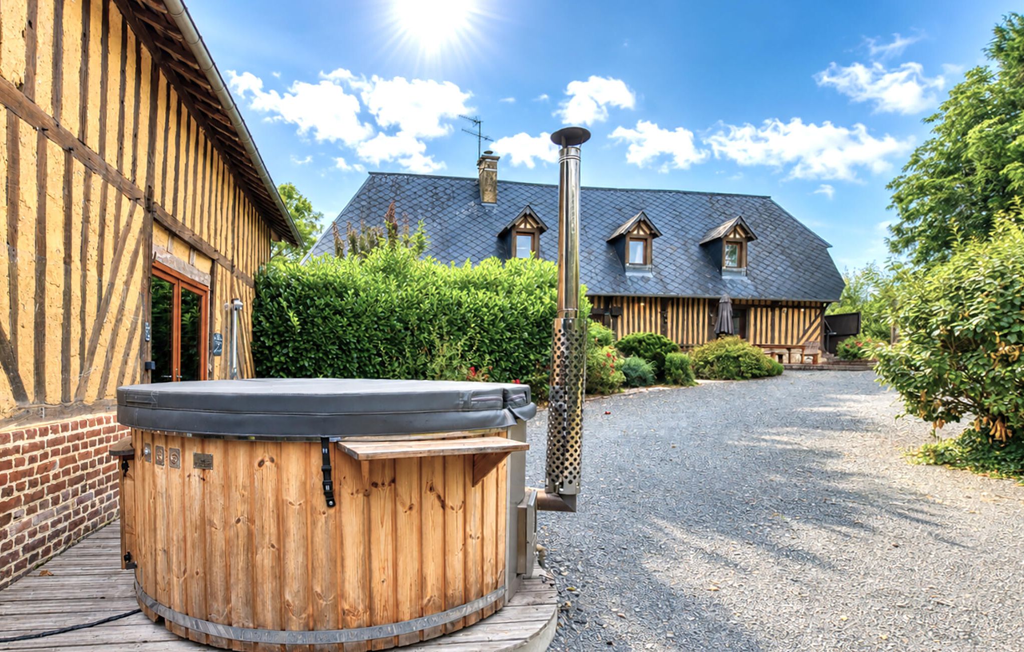 A wooden building with a hot tub in the foreground and a gravel driveway leading to a house with a dark roof and chimneys.